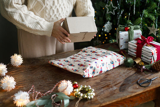 Woman in Cozy Knit Sweater Holding a Cardboard Gift Box over a Festive Wooden Table with Wrapping Supplies and Christmas Tree