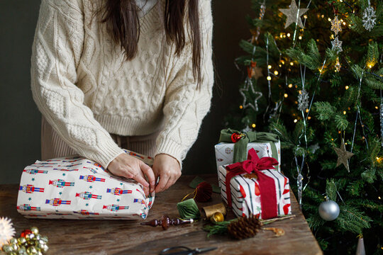Person Crafting Christmas Presents at a Wooden Table Surrounded by Nutcracker Paper, Ribbons, and a Glowing Tree. Handmade gift wrapping