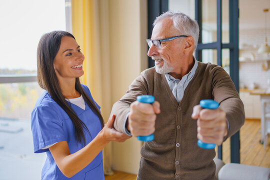 Young female nurse helping senior man exercise with dumbbells at home, caregiver supporting elderly patient during rehabilitation workout, home care service and recovery concept