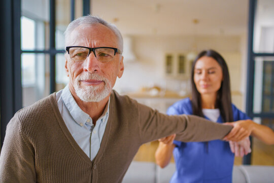 Young female nurse giving arm massage to elderly man at home, caregiver providing physical therapy support, rehabilitation treatment, home care service and healthcare concept