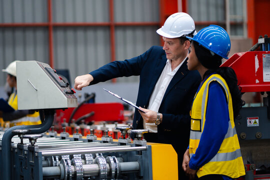 Two professional engineer,worker,technician use clipboard discuss work, walk in steel metal manufacture factory plant industry. Black African American man and woman wear hard hat check quality machine