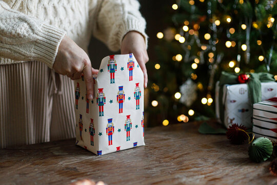 Woman hands folding nutcracker wrapping paper around Christmas gift box on rustic table, concept of winter DIY, creative present packaging, and festive seasonal preparation