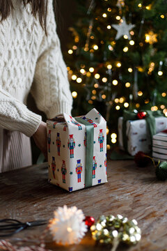 Woman wrapping Christmas present box on wooden table with decorated tree on background, concept of Christmas shopping, holiday spirit, family traditions, and handmade gift wrapping