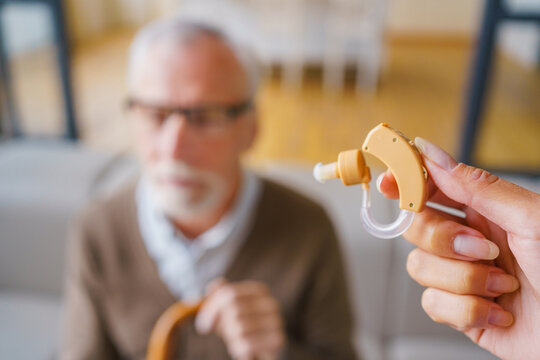 Focus on female hand holding hearing aid with blurred senior man in background, close up of assistive device for elderly care, hearing support, healthcare concept and rehabilitation assistance