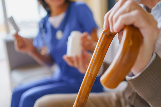 Female nurse giving pill jar to senior man at home, focus on hand with wooden cane , caregiver assisting elderly patient with medication, healthcare support and home care service concept