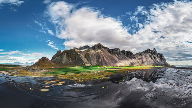 Aerial view of the dramatic Vestrahorn mountain peaks rising above the black sand beach and reflective tidal flats of Stokksnes in Iceland.