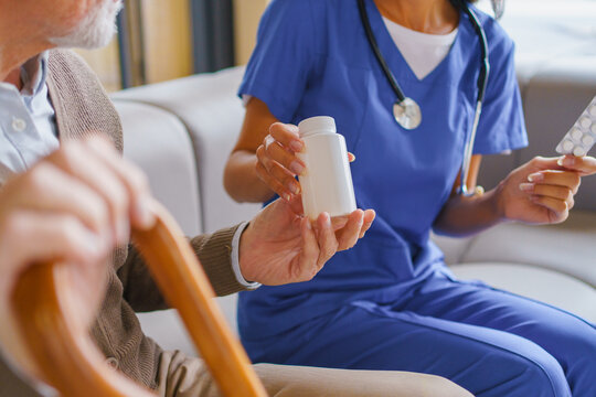 Female caregiver giving medicine jar to senior man at home, nurse assisting elderly patient with treatment, healthcare support and home care service concept