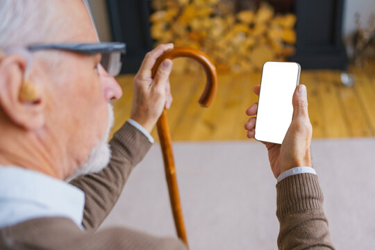 Rear view of senior man with wooden cane holding smartphone with black blank mock up screen, elderly male at home, mobile device display, copy space and senior lifestyle concept