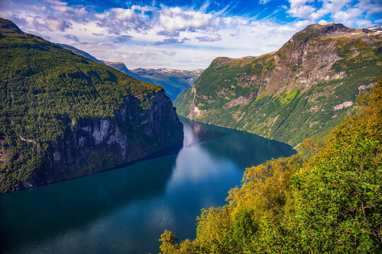 Scenic panorama of Geirangerfjord with steep green mountains, deep blue water, and dramatic clouds in summer, Norway.
