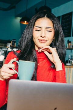 Entrepreneurial woman in a red sweater enjoys coffee while working on a laptop in a cozy cafe setting