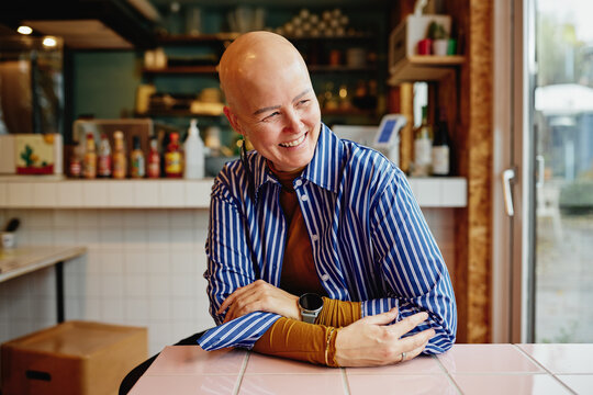 Smiling bald woman in striped shirt sits at a cafe counter, relaxed and friendly, enjoying a warm moment