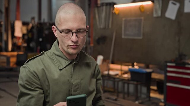 Young adult Caucasian male worker sitting in workshop, drinking from thermos and using smartphone. Video is suitable for industrial break time, blue-collar lifestyle, and mobile app promotions