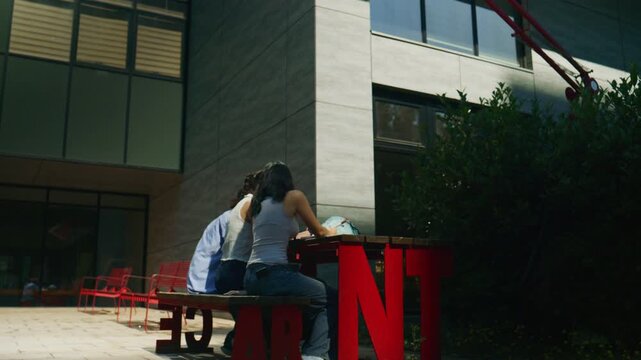 Three high school students are diligently working on their assignments at an outdoor table. They are collaborating on a study project in a bright, modern academic setting.