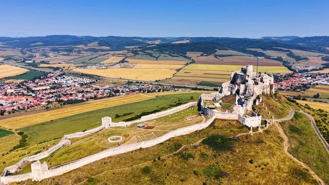 Wide aerial shot of Spis Castle with nearby town. Medieval fortress on high hill with surrounding stone walls and adjacent village in valley, clear sunny day.