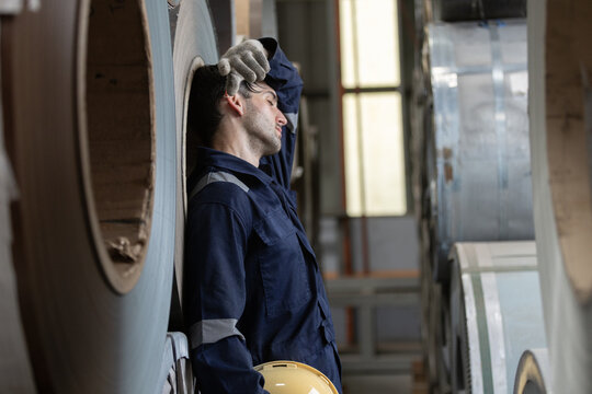 Tired factory worker wipes sweat while standing beside large steel coils in an industrial facility, representing workplace fatigue, heat stress, manufacturing labor, safety, occupational health.