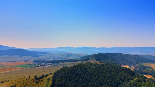 Mountainous landscape with forested hills and valley. Forested hill sits in the foreground with a wide valley and distant mountain ranges under a clear sky.