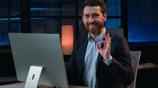 Positive Caucasian middle-aged business man with computer showing okay gesture at night dark office adult 40s businessman male executive work online demonstrate ok sign happy web job task at evening
