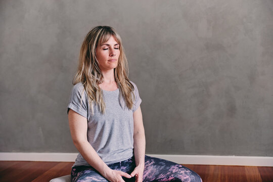 A woman is sitting  in yoga class, with cross-legged on the floor with her eyes closed, practicing breathwork and meditation.