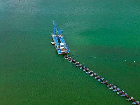 Aerial view of a blue sand mining dredger operating in a calm green lake with a long floating pipeline extending across the water, Poland.