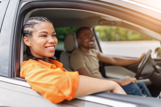 Cheerful young African American woman resting her arm out the car window and smiling, with man driving blurred in background during a relaxed sunny road trip