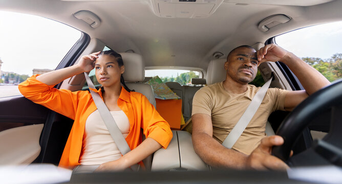 Tired young African American couple sitting in car with tense expressions, both rubbing their foreheads in stress, showing exhaustion during a long road trip
