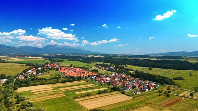 Aerial view of countryside village and strip farmland, Slovakia. High angle panorama of yellow crop fields, red roofs, dense woodland, and mountain peaks under blue sky.