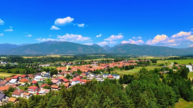 Summer mountain view of town. A small town is located near mountains on a sunny day with scattered clouds. Houses are visible among green trees.