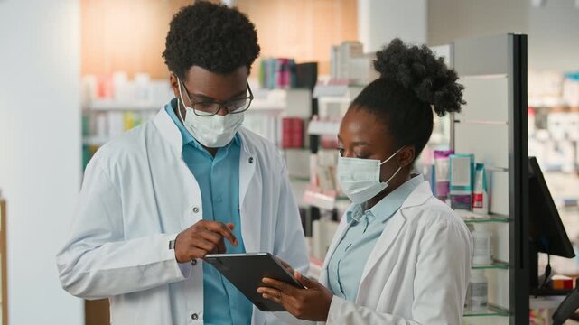 Two African American doctors pharmacists wearing medical masks discussing digital tablet health medicine data in pharmacy therapists talking looking in device in drugstore clinic coronavirus pandemic