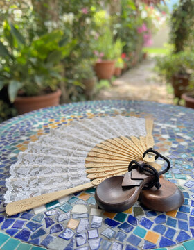 A folded lace fan and a pair of wooden castanets lie on a colorful mosaic tile table in a garden.