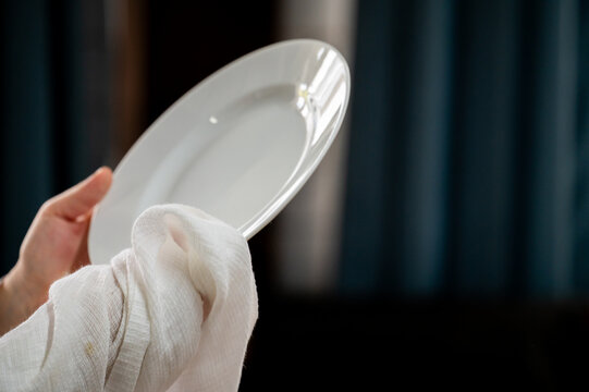 Close-up of a person's hands wiping a clean white ceramic plate with a white cotton cloth towel, standing indoors against a blurred dark blue curtain background.