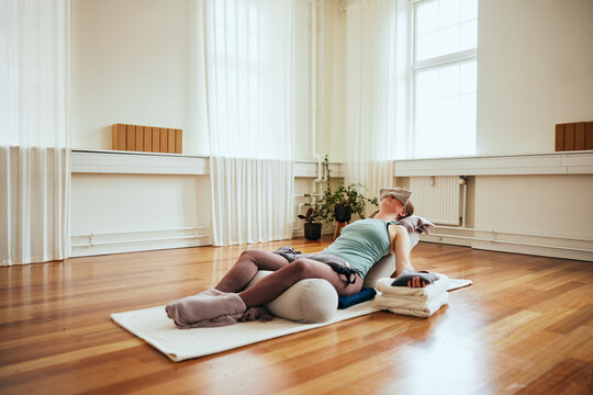 Woman doing restorative yoga using bolsters. She is lying on the floor on a mat with her arms outstretched to each side