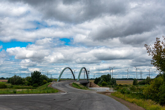 Wide view of modern steel arch bridge with curving road and wind turbines on horizon under cloudy sky in Wustermark Germany with copy space