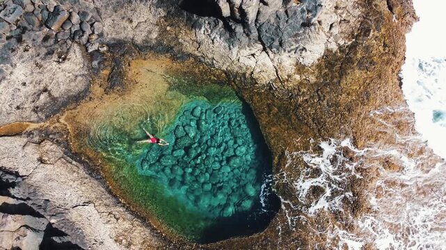 Aerial top down view of woman relaxing in a turquoise tidal pool surrounded by volcanic rocks