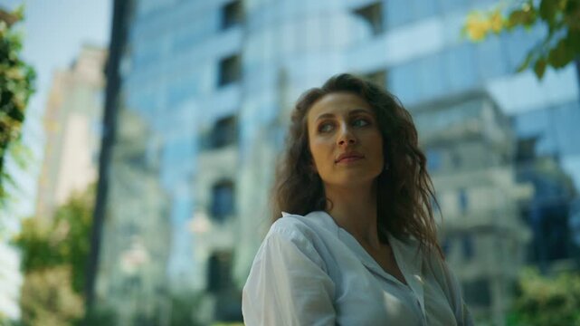 A confident businesswoman with curly hair stands thoughtfully in front of a modern glass building. She observes the urban environment, embodying a contemplative and independent spirit.