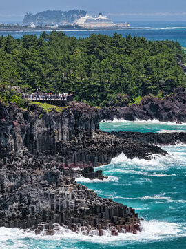 Aerial view of the basaltic columnar joints of the Jusangjeolli Cliffs with turquoise waves, dark volcanic rocks, and lush forest, Jeju Island, South Korea.