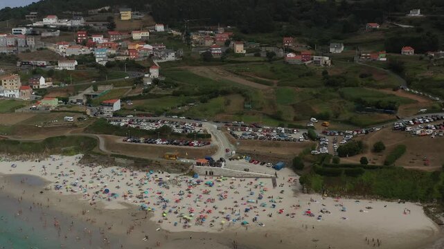 Aerial view of the coastal town of Caion with its busy beach, colorful umbrellas, and large parking areas in Caion, A Coruna, Spain.