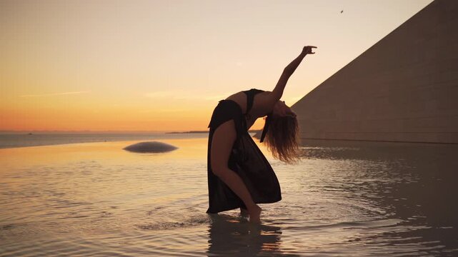 Young woman dancing in shallow water during sunset, performing sensual expressive freestyle with graceful poses showing emotion and freedom