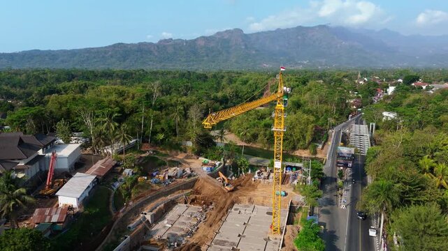 Aerial view of a construction site featuring a tall yellow tower crane, heavy machinery, and ongoing building work surrounded by lush greenery in Mungkid, Central Java, Indonesia.