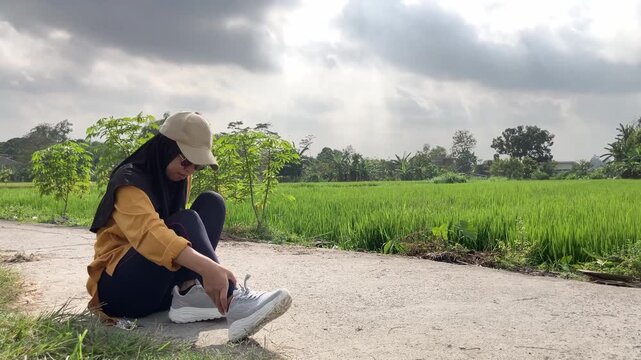 Slow motion video of a young Muslim woman in a hijab standing up and limping after suffering a sprained ankle. She walks with difficulty on a path next to a green rice field. Sports concept.