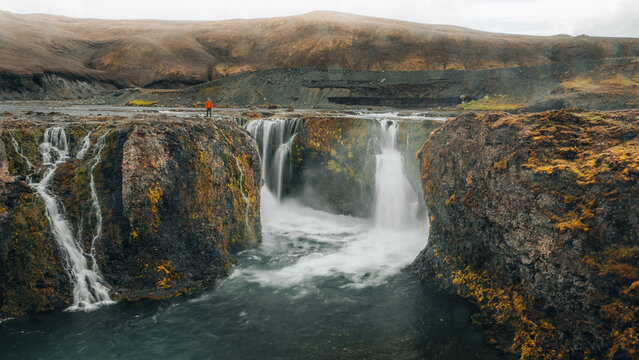 Hiker standing above a powerful waterfall in Asahreppur, Iceland, surrounded by rugged volcanic rock and rushing water, capturing the raw and dramatic beauty of Icelandic wilderness.