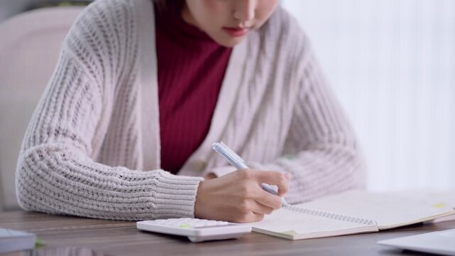Young Asian student writing notes in notebook during focused study session at home desk with copy space