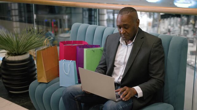 Busy African American mature business man typing laptop in shopping mall with bags purchases middle-aged businessman concentrated male executive working distant with computer ordering e-commerce goods