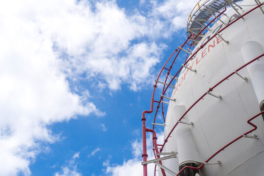 Low angle view of a large white spherical propylene storage tank with red piping and ladder against a blue sky with white clouds at a petrochemical industrial plant.