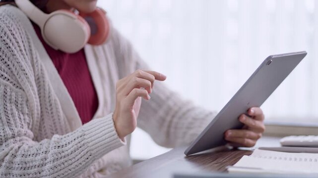 Adult Asian woman using digital tablet while listening to headphones at home workspace with copy space