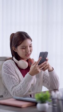 Young Asian woman smiling while using smartphone with pink headphones at home desk with copy space