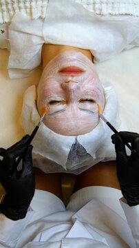 Professional beautician applying cleansing mask on female client's face. Close-up of female face during youth restoration and facial rejuvenation procedure.