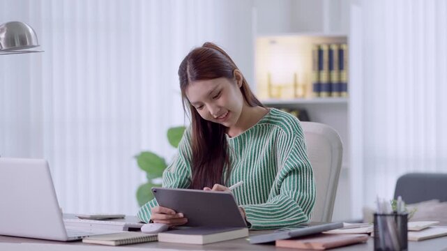 Young Asian professional using tablet and laptop for remote work at bright home office desk