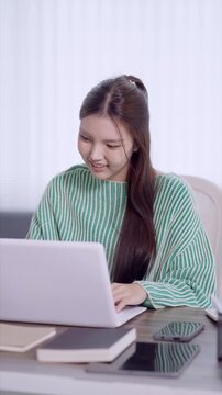 Young Asian student working on laptop computer at home desk with books and smartphone