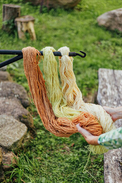 Skeins of wool yarn in warm brown, pale yellow and cream tones hang from a metal rod while a person handles the fibers outdoors in a rustic natural setting