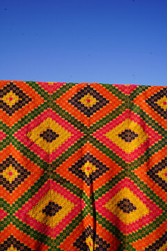 Vertical shot of vibrant handwoven kilim rug with colorful diamond patterns hanging outdoors under a clear blue sky. Traditional Greek folk art textile display bathed in direct sunlight.